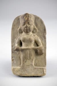 A carved stone sculpture depicting a standing figure wearing a headdress and large earrings, with an expressionless face. The figure holds a flat object and appears to have detailed carvings of jewelry and attire. The background of the sculpture is smooth.