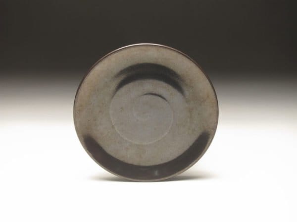 An overhead view of the Pastry Plate by Zane Wilcox with a minimalist design. The plate has a matte gray finish with darker, almost black, circular brushed patterns near the rim and center. The background is softly lit, creating a gradient from light to dark.