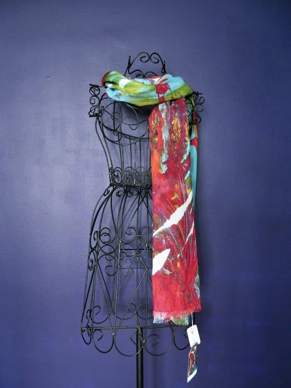 A **Something About You Scarf by Kay Jay Studio** with a vibrant mix of red, green, and blue hues is draped over the shoulders of an ornate black wire dress form against a dark blue wall. The scarf features abstract patterns and a white price tag is visible hanging from it.