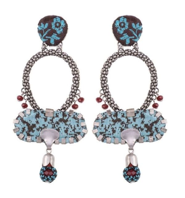 A pair of Earrings by Ayala Bar featuring intricate blue and brown embroidery on circular fabric, silver chains forming oval shapes with red beads, and decorated with blue and brown beads and silver accents hanging from a broad, patterned fabric at the bottom.