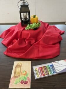 A metal lantern, plastic grapes, a pepper, and cherries arranged on red fabric for a still life reference while drawing