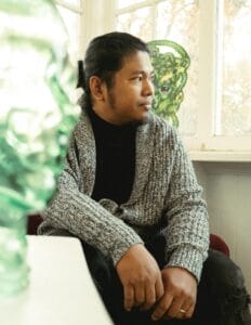 A person sitting in a room with natural light, wearing a gray cardigan and black top. They have long hair tied back and appear to be looking out of the window. A green glass sculpture is in the foreground.