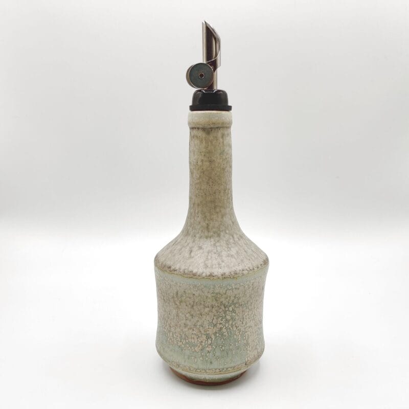 A tall, textured ceramic bottle with a narrow neck and speckled glaze features a metal bottle pourer inserted into the top, styled in a minimalist Bazaart 2025 aesthetic and set against a plain white background.