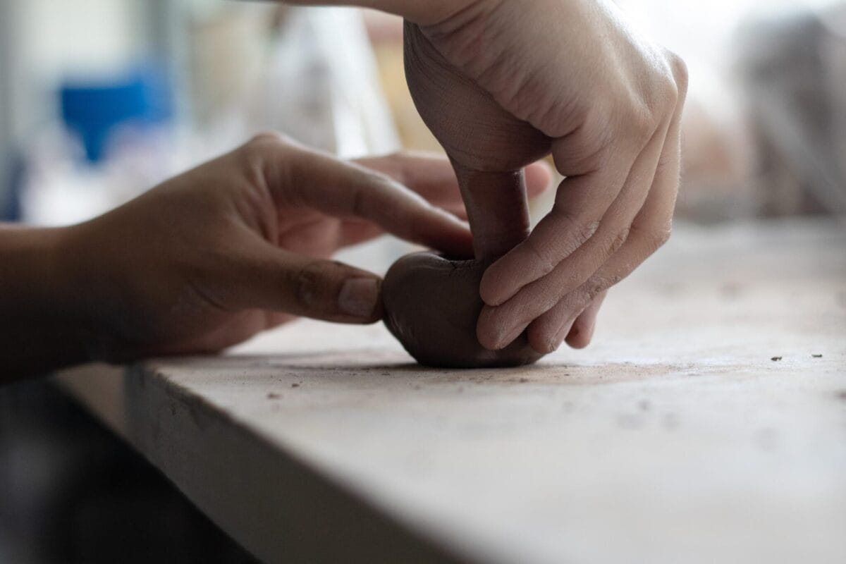 Close-up of hands shaping a small piece of clay on a flat surface. The person is using their fingers to press and mold the clay into a pinch pot, demonstrating a pottery or sculpting activity. The background is softly blurred, emphasizing the hands and clay in this portrait.