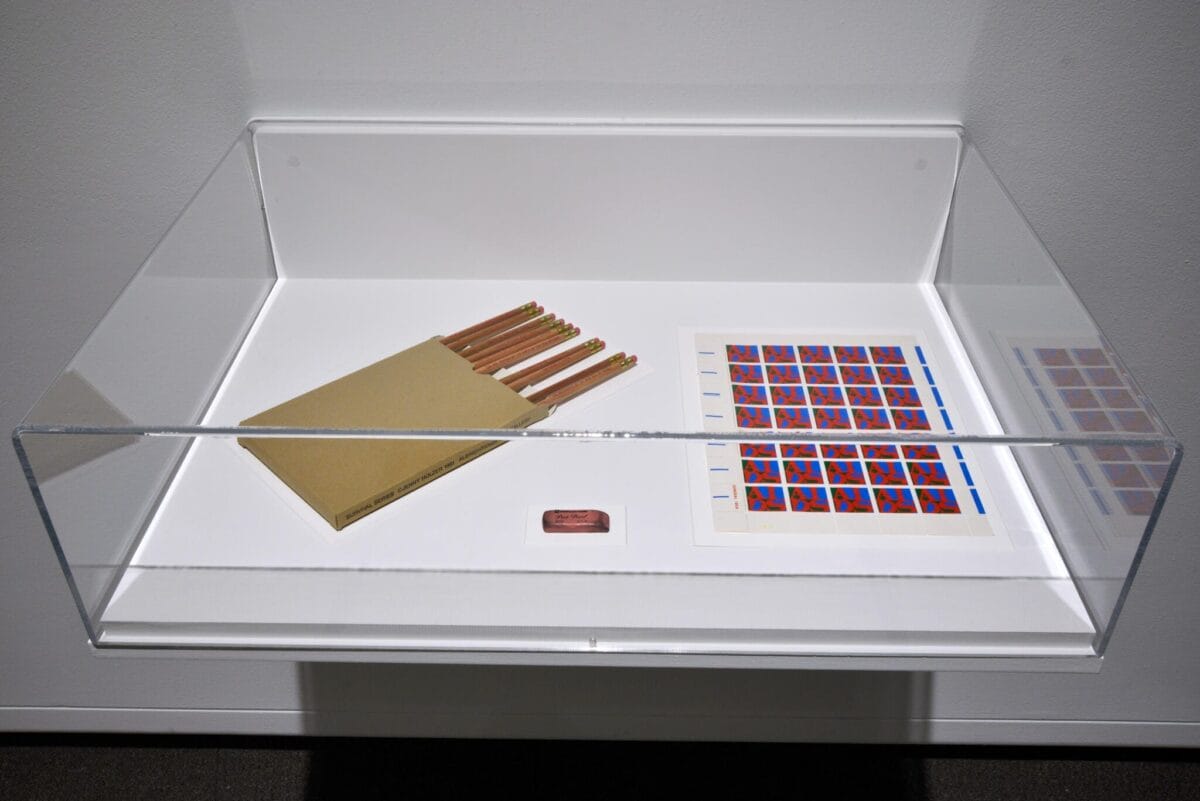 A display case, as thick as thieves with secrets, holds a beige folder filled with pencils, a copper plaque, and a sheet showcasing a vibrant grid of stamps. The case stands proudly mounted on a pristine white wall.
