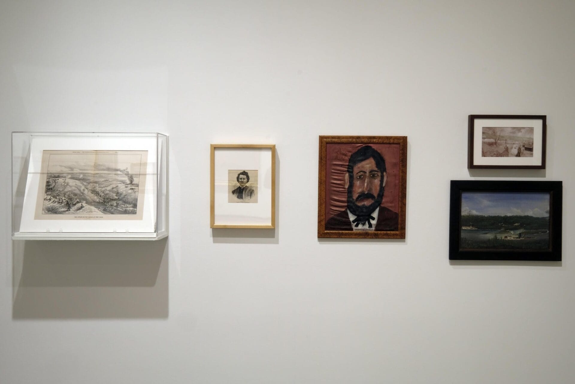 A white wall displays five framed artworks: a detailed landscape in a glass case, a small portrait sketch, a larger painting of a man with a mustache and beard, followed by two more small landscapes in varying frame styles and colors.