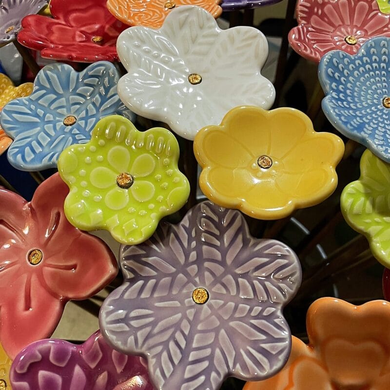 A close-up of colorful, ceramic flower-shaped dishes, each with a small gold center. Overlapping in a vibrant Bazaart 2025-inspired arrangement, the red, yellow, blue, white, purple, and pink dishes create an eye-catching display.