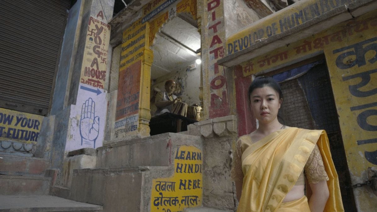 A woman in a yellow sari stands in front of an alleyway adorned with colorful signs promoting yoga, meditation, and learning Hindi. In the background, an elderly person sits in a small, open shop or temple. The scene appears to be in an urban setting.