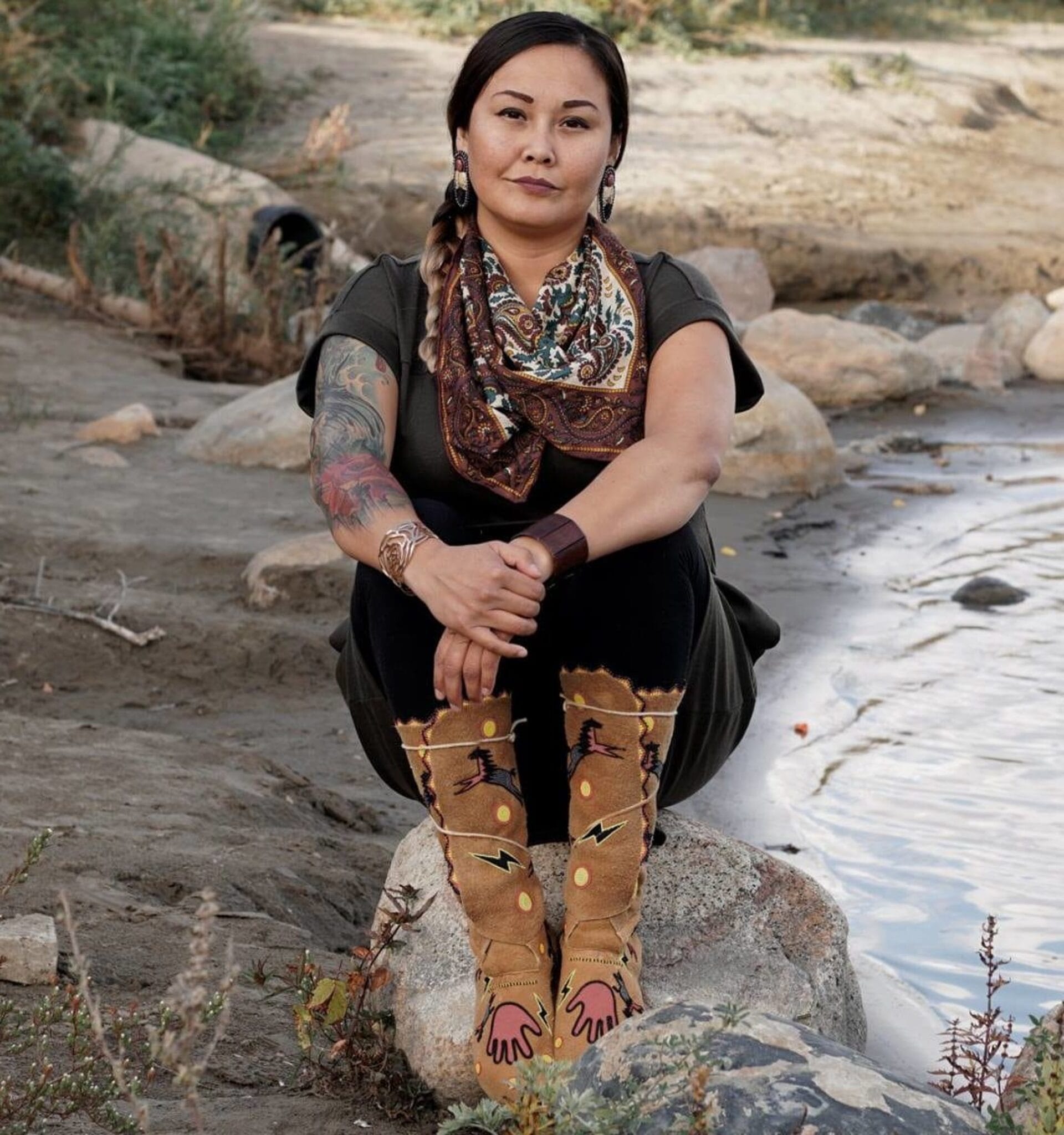 A person with long dark hair sits on a rock near a body of water, wearing a dark green dress, a patterned scarf, and ornamental boots with decorative patterns. They have tattoos on their arms and large earrings. The natural background, featuring rocks and vegetation, contrasts beautifully with the free-spirited scene.