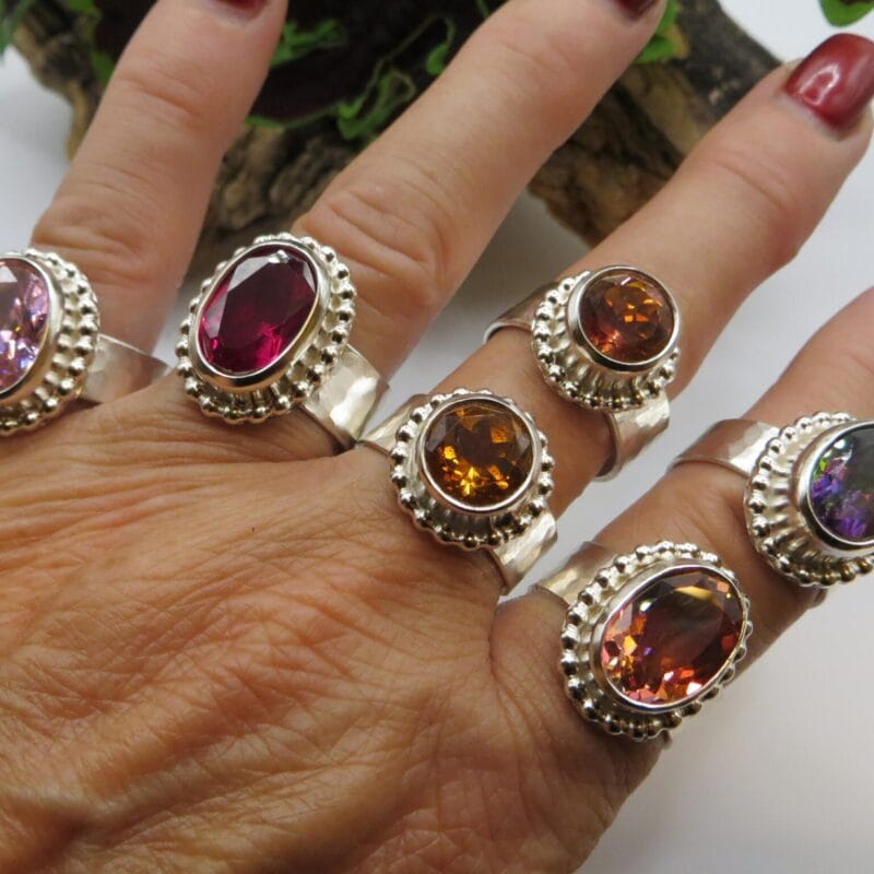 A hand adorned with five silver rings, each showcasing large, colorful oval gemstones in purple, pink, red, orange, and multicolor hues, evokes the festive spirit of Holiday Bazaart 2024. Dark red nails contrast beautifully against a lush backdrop of greenery.