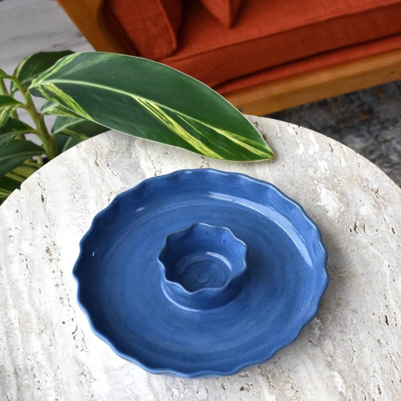A blue, wavy-edged ceramic chip and dip platter, inspired by Bazaart 2025 trends, sits on a round, light-colored stone table with a green leafy plant nearby and an orange couch in the background.