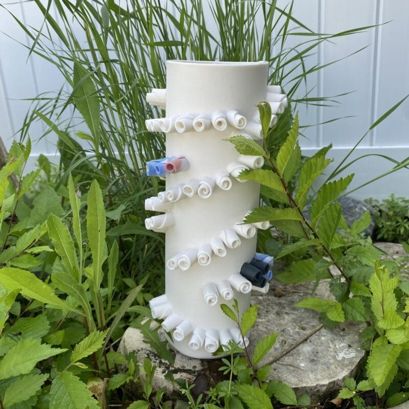 A white PVC pipe stands upright outdoors, surrounded by green plants and rocks. Spiraled white cones with one red and one blue connector embellish its surface, reminiscent of a modern sculpture for Holiday Bazaart 2024. Tall grass waves in the background against a sleek white fence.