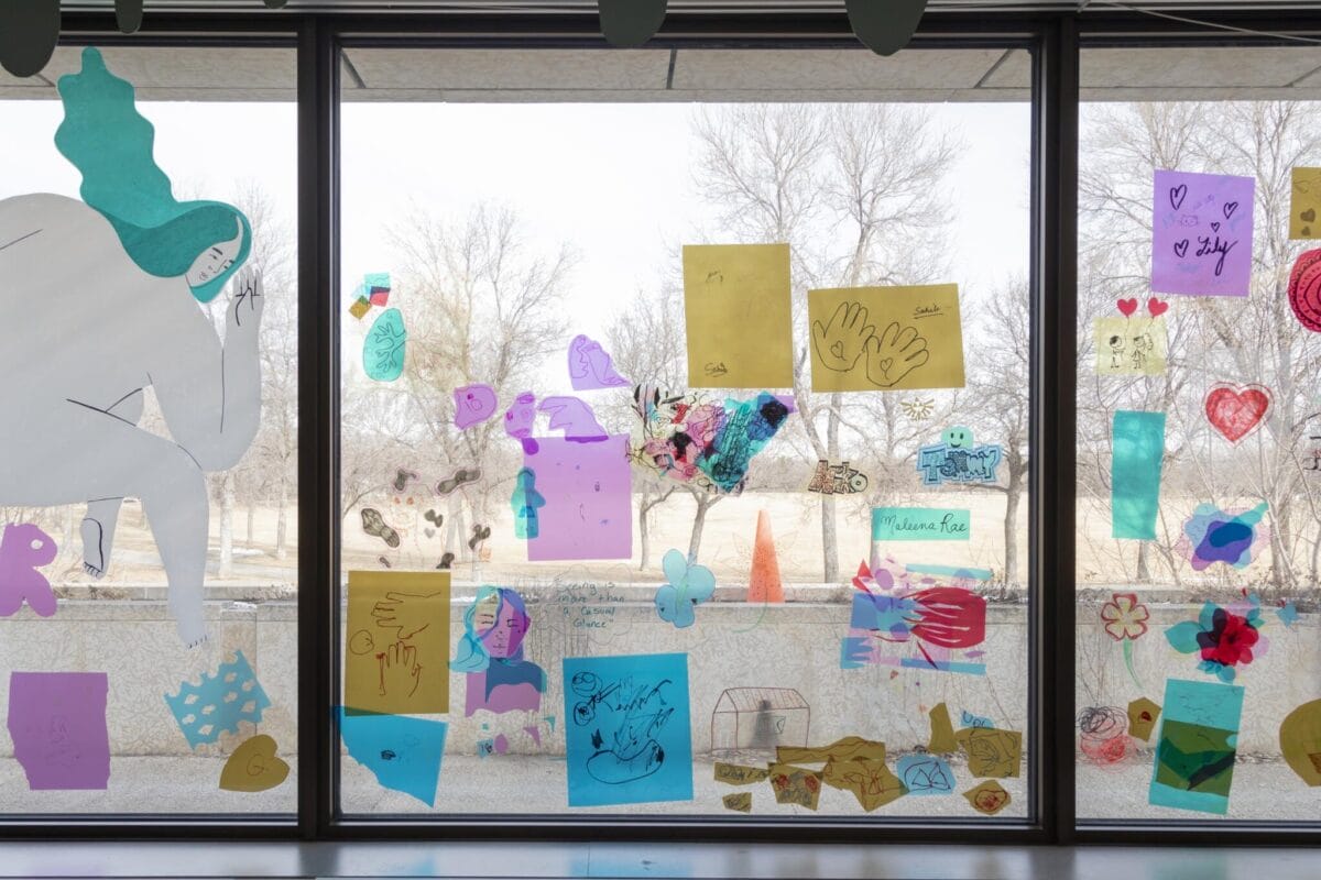 A large window displays various colorful drawings and cut-out paper shapes, including hearts, hands, and animals. The background shows leafless trees and an outdoor scene through the window. Some doodles are taped, creating a vibrant and creative montage.
