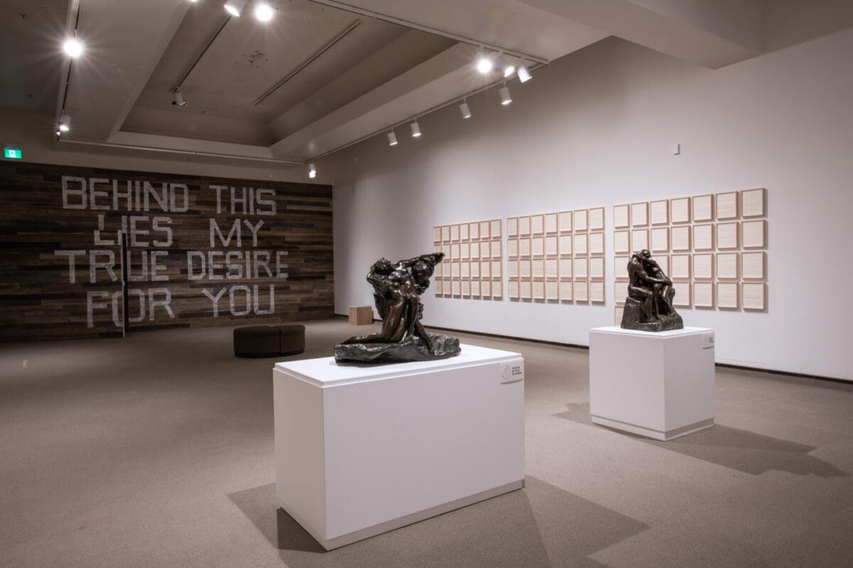 Art gallery with two sculptures on white pedestals. A wooden wall displays the text