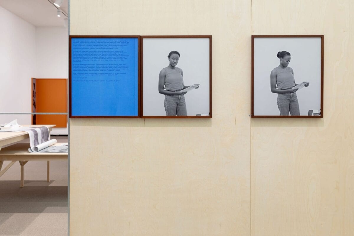 Two black-and-white portraits of a woman holding a book are displayed on a light wooden wall in a gallery. The left frame also includes a blue text panel. A table on the left side of the room holds documents or artwork. The background includes additional displays.