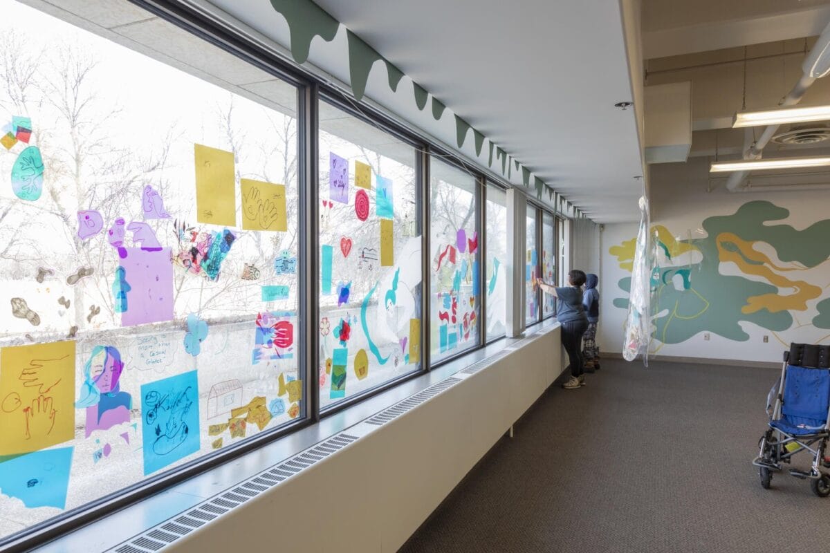 A person adds colorful drawings to a large window in a bright, spacious room. The window is adorned with various vibrant art pieces, while a wheelchair is seen to the right. The room has abstract wall art and plenty of natural light streaming in.