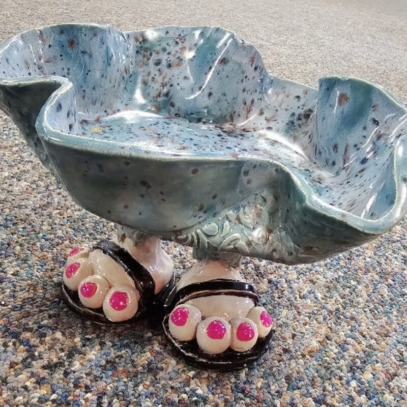 A wavy, blue ceramic bowl with speckles is supported by two sculpted feet wearing black sandals with pink toenails, set on a textured, multicolored carpet—a playful Bazaart 2025 creation.