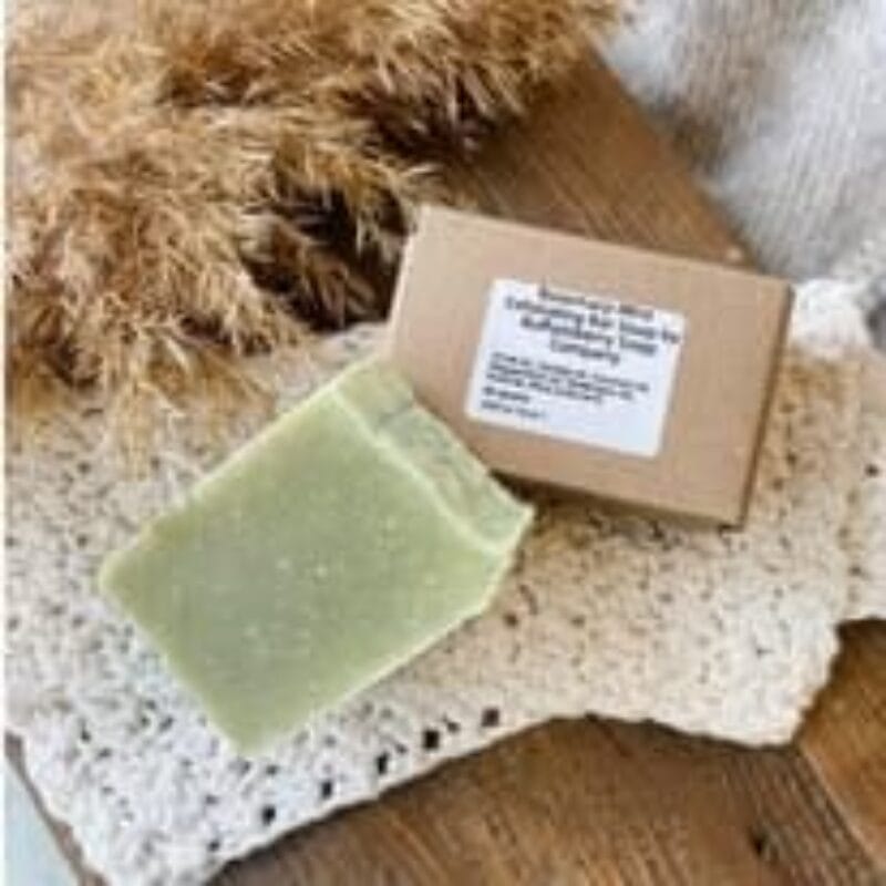 A green soap bar and a brown box with a label sit on a wooden surface. The soap rests on a white crocheted cloth, with dried grass in the background, channeling a rustic and natural vibe reminiscent of Bazaart 2025's serene aesthetics.
