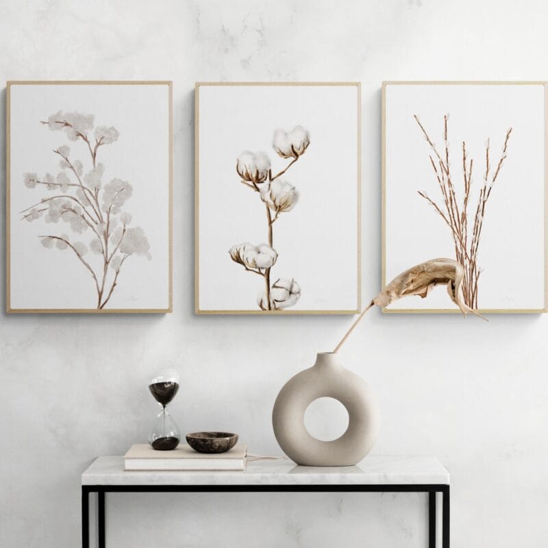 A minimalist room with a small marble table is the perfect setting for your Holiday Bazaart 2024 finds. On the table, a modern vase with a dried leaf, a round bowl, and an hourglass sit elegantly beneath three framed botanical art prints adorning the light wall.