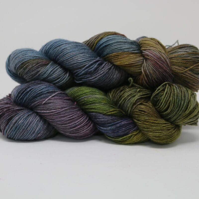 Three skeins of yarn in muted holiday colors, including shades of blue, green, purple, and brown, are artfully arranged against a plain white background. Perfect for your upcoming Holiday Bazaart 2024 projects, the yarn appears soft with a subtle sheen, showcasing harmonious tones.