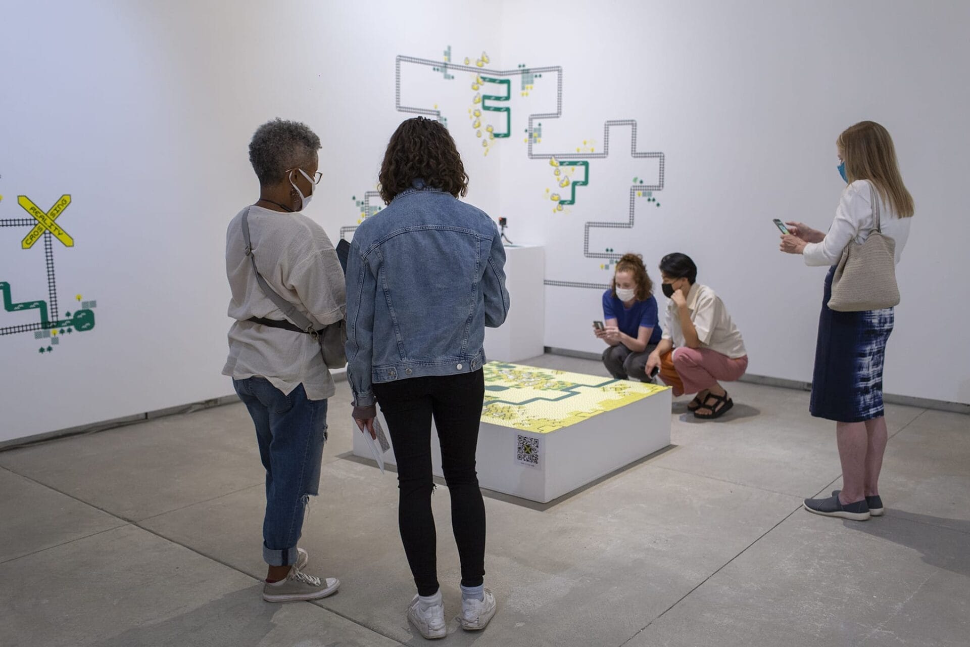 People observe an art exhibit featuring geometric designs on a gallery floor and wall. Some visitors take photos while others view the artwork closely. Most are wearing casual clothing and masks.
