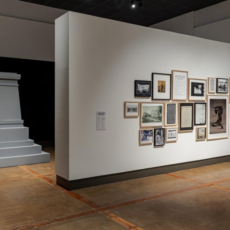 A gallery room features a pedestal structure and a wall decorated with an array of framed photos, documents, and newspaper clippings. The artworks are neatly arranged in various sizes and styles on the white wall. The room has a minimalist design and polished concrete floors.