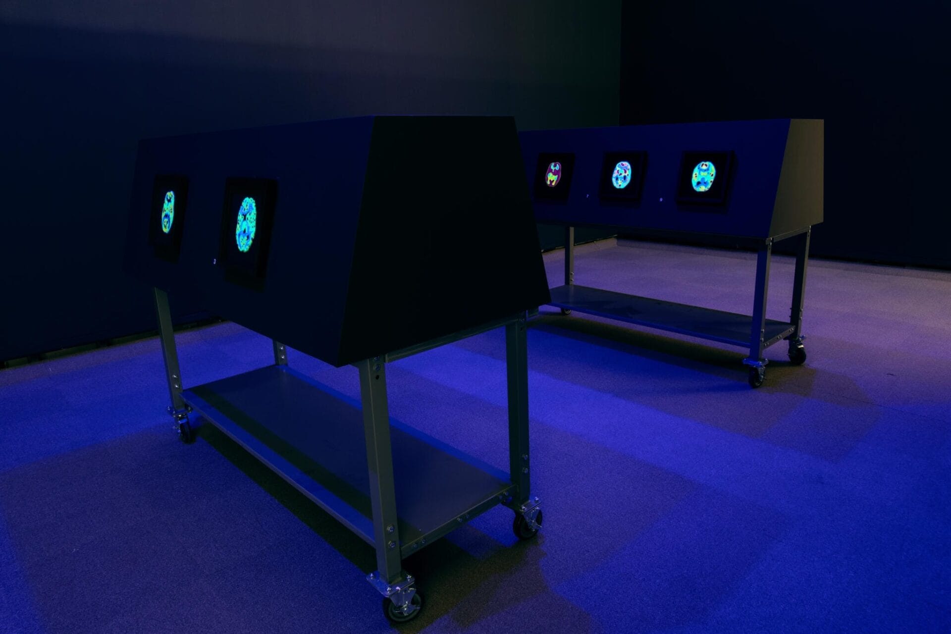 Two dark rectangular installations on wheels display digital screens with colorful geometric patterns, so small we don't even think about it. The room's dim lighting casts a blue hue, enhancing the modern and futuristic atmosphere.