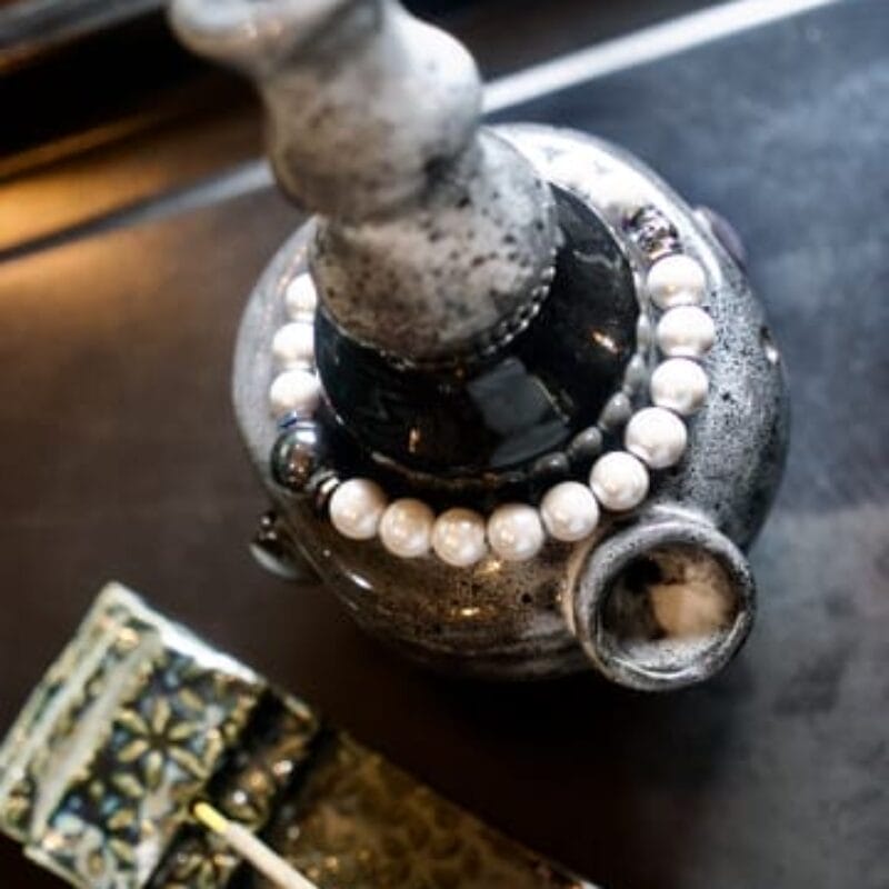 A close-up of a ceramic vase with a string of white pearls draped around its neck, placed on a dark surface next to a textured green ceramic object, captured in the refined style of Bazaart 2025.