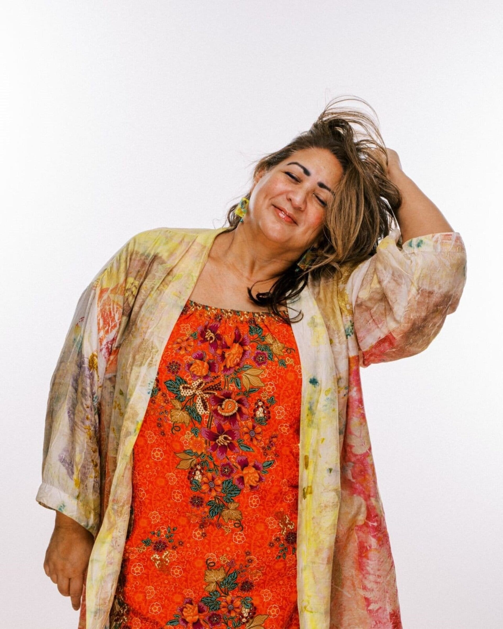 A woman wearing a brightly colored floral dress and a patterned overcoat stands against a plain white background. She has a content expression with her eyes closed and one hand playfully running through her tousled hair.