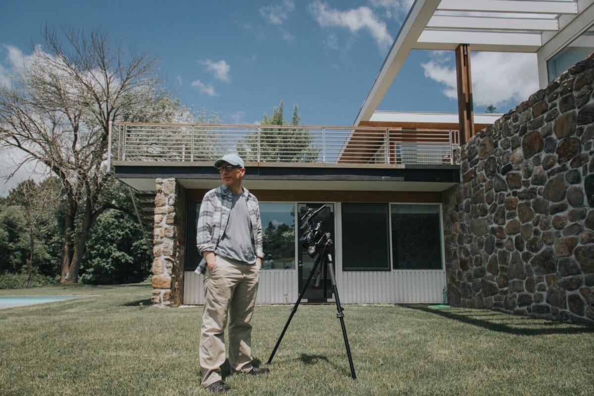 A person in casual attire stands next to a camera on a tripod in front of a modern house with a stone facade. The house is set against a backdrop of a clear blue sky and trees.