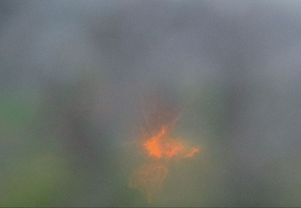 A photograph depicting a soft, misty background with an abstract, glowing orange and yellow light at the center. The light has a starburst pattern, contrasting with the diffuse, muted colors surrounding it.