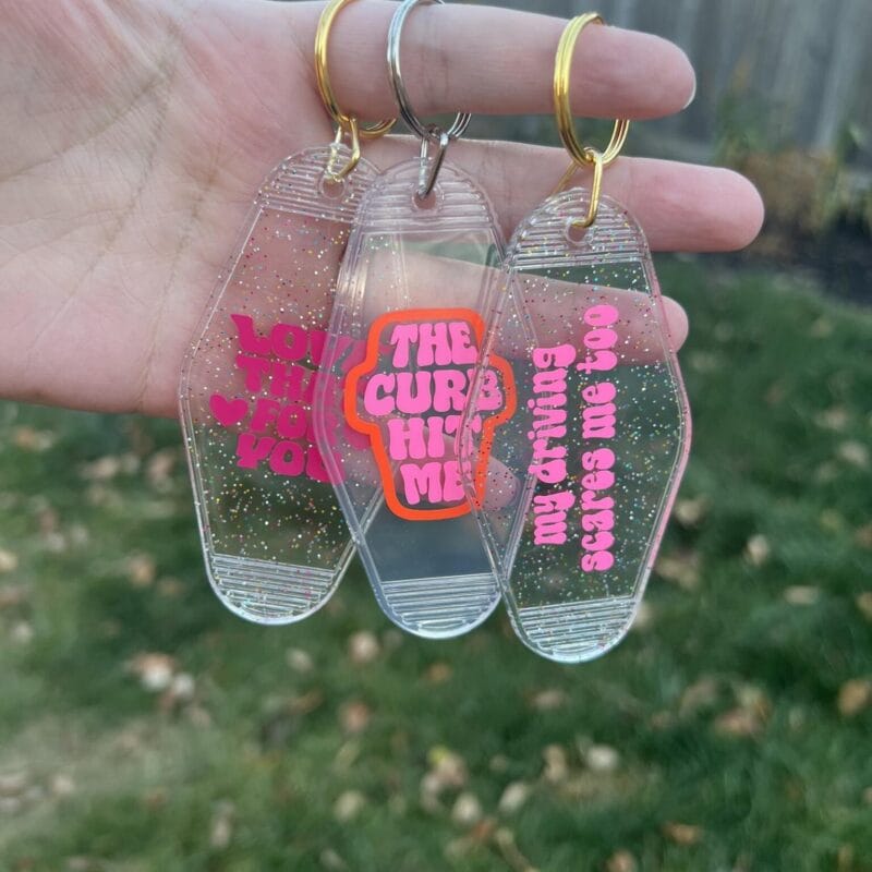 A hand holds three clear coffin-shaped keychains with pink and orange text that reads