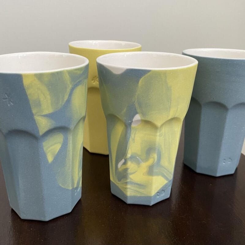 Four tall, faceted cups sit on a dark wooden surface. Two are solid blue, one is yellow, and two showcase blue and yellow marbled patterns, creating a modern Bazaart 2025 vibe against the plain, light-colored background.