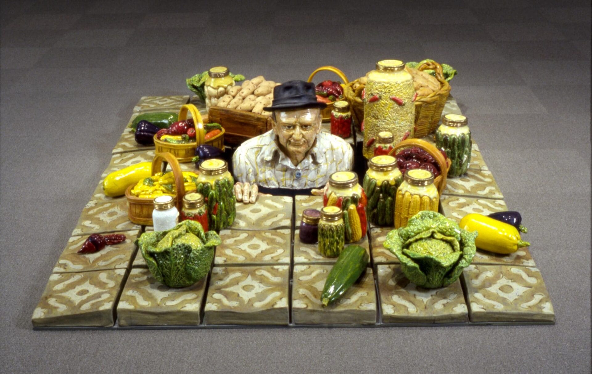 A sculpture of a man wearing a hat and a patterned shirt is emerging from a tiled ground surrounded by various baskets and jars filled with vegetables, including peppers, lettuce, cucumbers, and squash in vibrant colors.