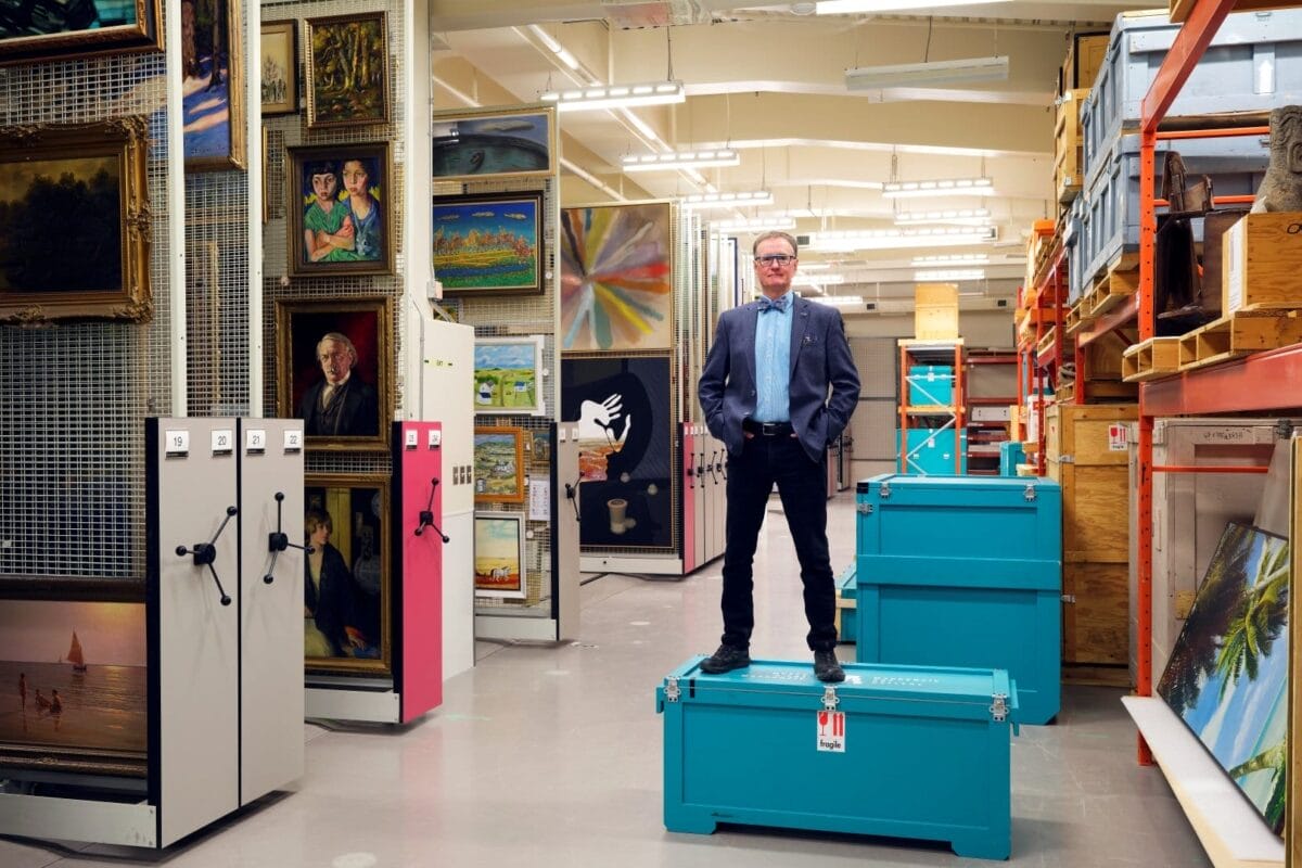 A person wearing a blazer and glasses stands confidently on a teal storage trunk in an organized storage area filled with paintings and other artworks. Whispers from the Vault, the space features neatly arranged shelving and racks filled with various framed art pieces and storage boxes.