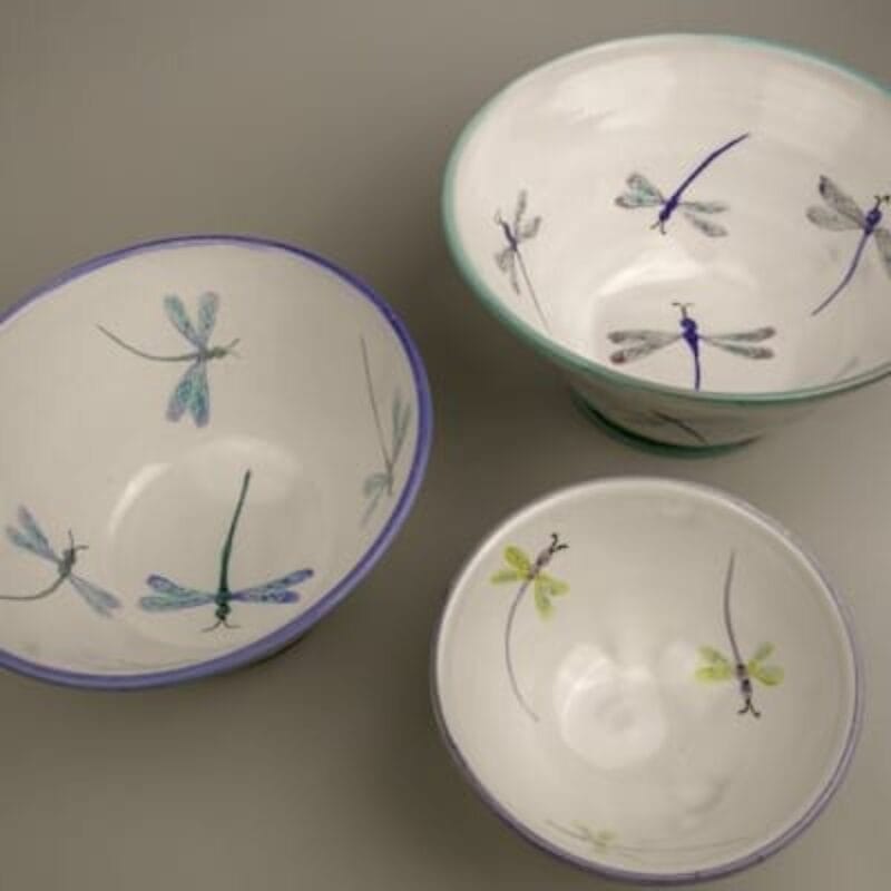 Three ceramic bowls with dragonfly designs are showcased at Holiday Bazaart 2024 on a grey surface. Each bowl boasts a unique pattern, featuring blue, green, and purple dragonflies. The bowls vary in size and have colorful rims matching the dragonfly accents.