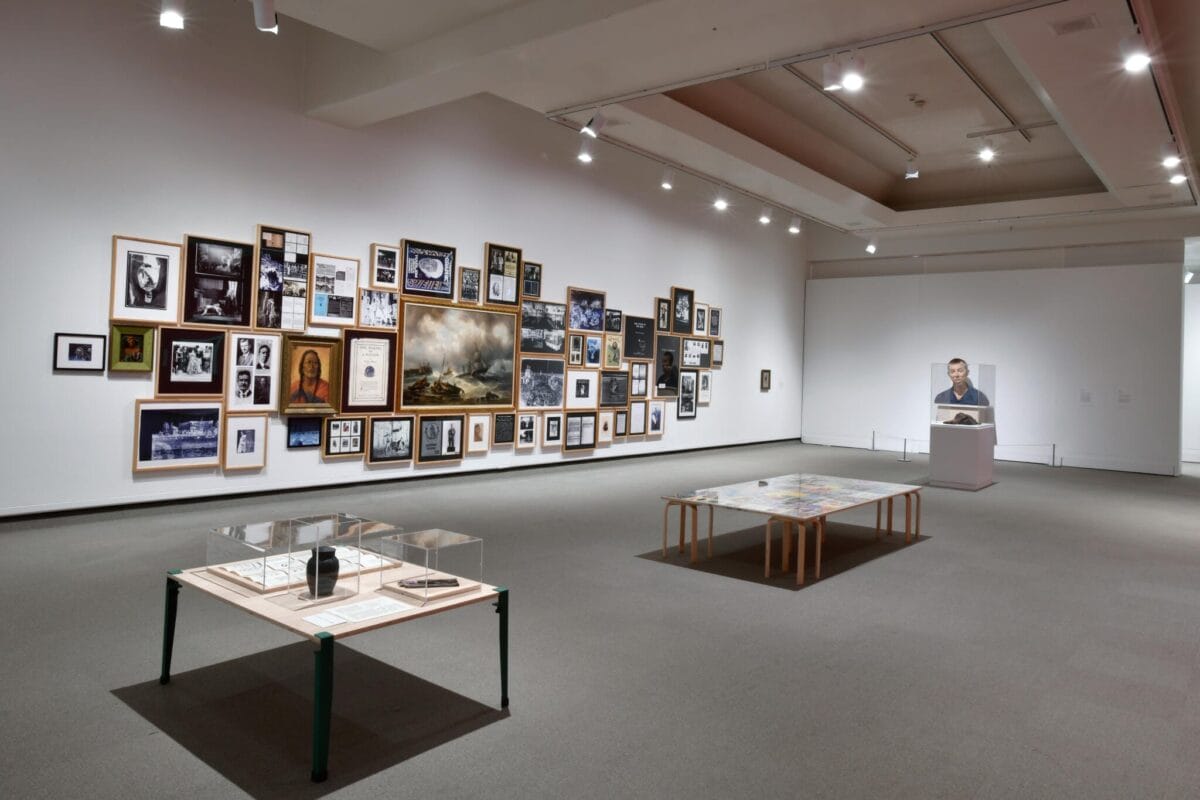A spacious art gallery with white walls, featuring a large photo collage on the left wall reminiscent of