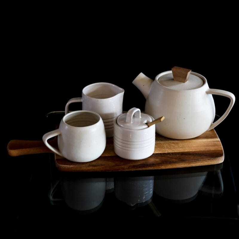 A white ceramic teapot, two matching cups, and a sugar bowl with a wooden spoon sit on a wooden serving board against a black background, styled for Bazaart 2025.