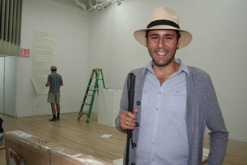 A smiling man in a light gray button-up shirt, light gray cardigan, and beige hat stands in a room with a wooden floor. He holds a black object in his hand and a green step ladder and another person with gray shorts and shirt can be seen in the background.
