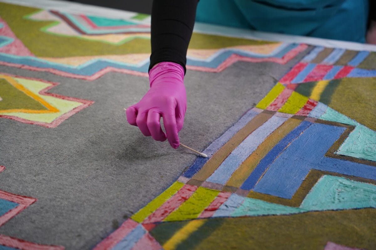 Une personne portant des gants roses et un tablier bleu sarcelle utilise un petit outil pour ajouter des détails à une peinture géométrique vibrante. L'œuvre présente des couleurs vives et des motifs complexes sur une surface texturée.
