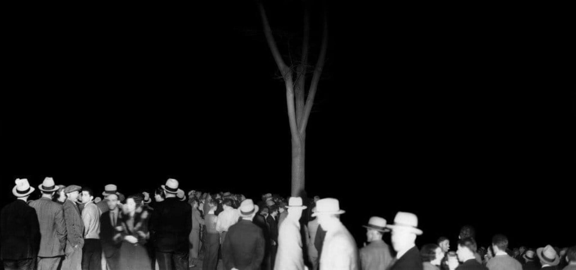 A black-and-white photo shows a crowd of people gathered under a tree at night. Many of them are wearing hats and appear to be engaged in conversation. The background is completely dark, with no other visible details.