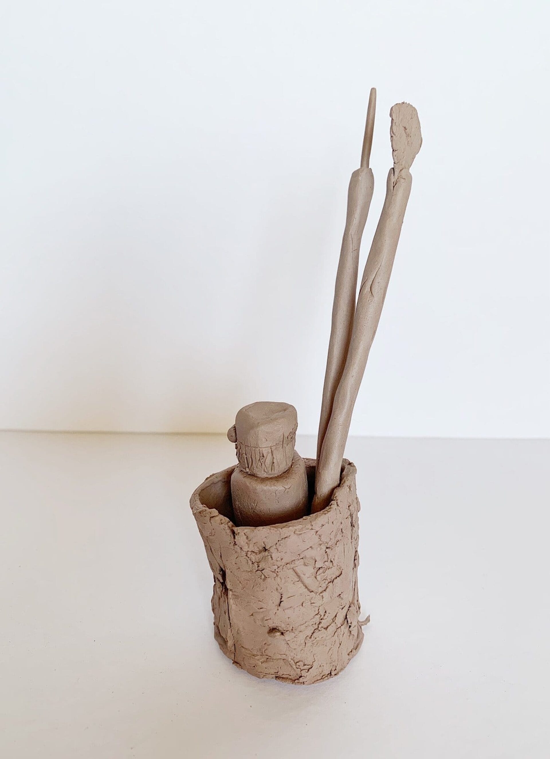 A simple clay sculpture depicts a cylindrical pot holding two elongated, vertical tools, resembling a paintbrush and sculpting tool. The pot and tools have a rough, textured finish, and the background is plain white.