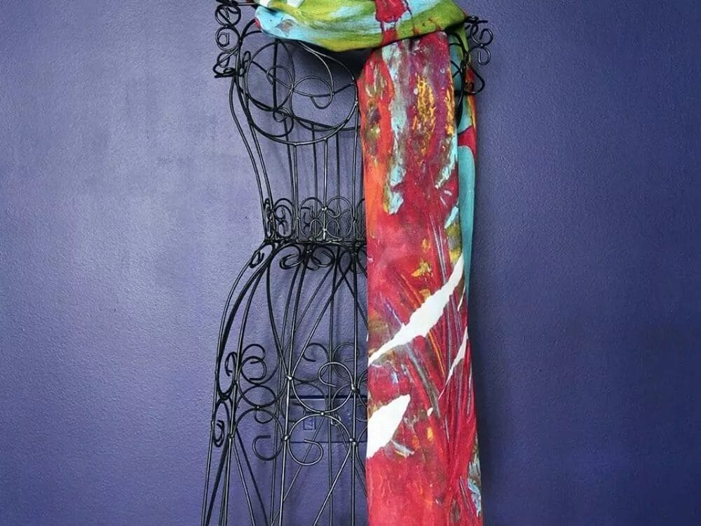 A **Something About You Scarf by Kay Jay Studio** with a vibrant mix of red, green, and blue hues is draped over the shoulders of an ornate black wire dress form against a dark blue wall. The scarf features abstract patterns and a white price tag is visible hanging from it.