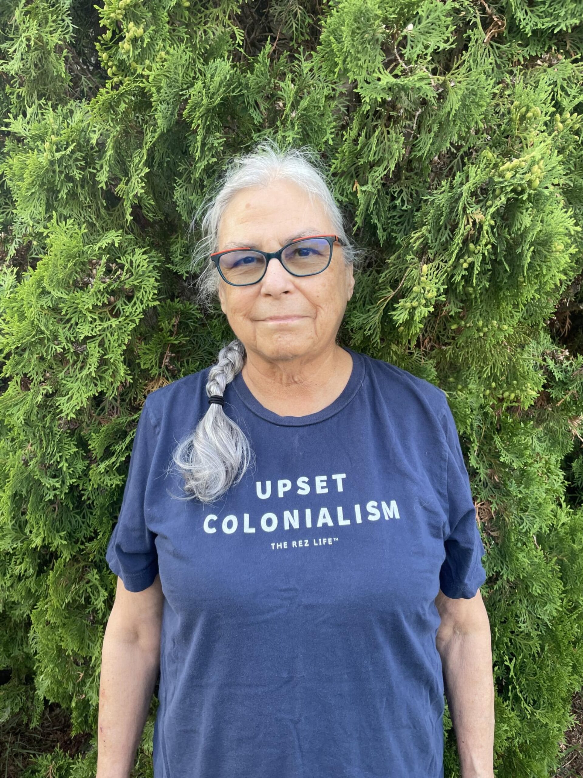 Une femme âgée aux longs cheveux gris tressés sur une épaule se tient devant un feuillage vert dense. Elle porte des lunettes et un T-shirt bleu sur lequel on peut lire « UPSET COLONIALISM » et « THE REZ LIFE ».