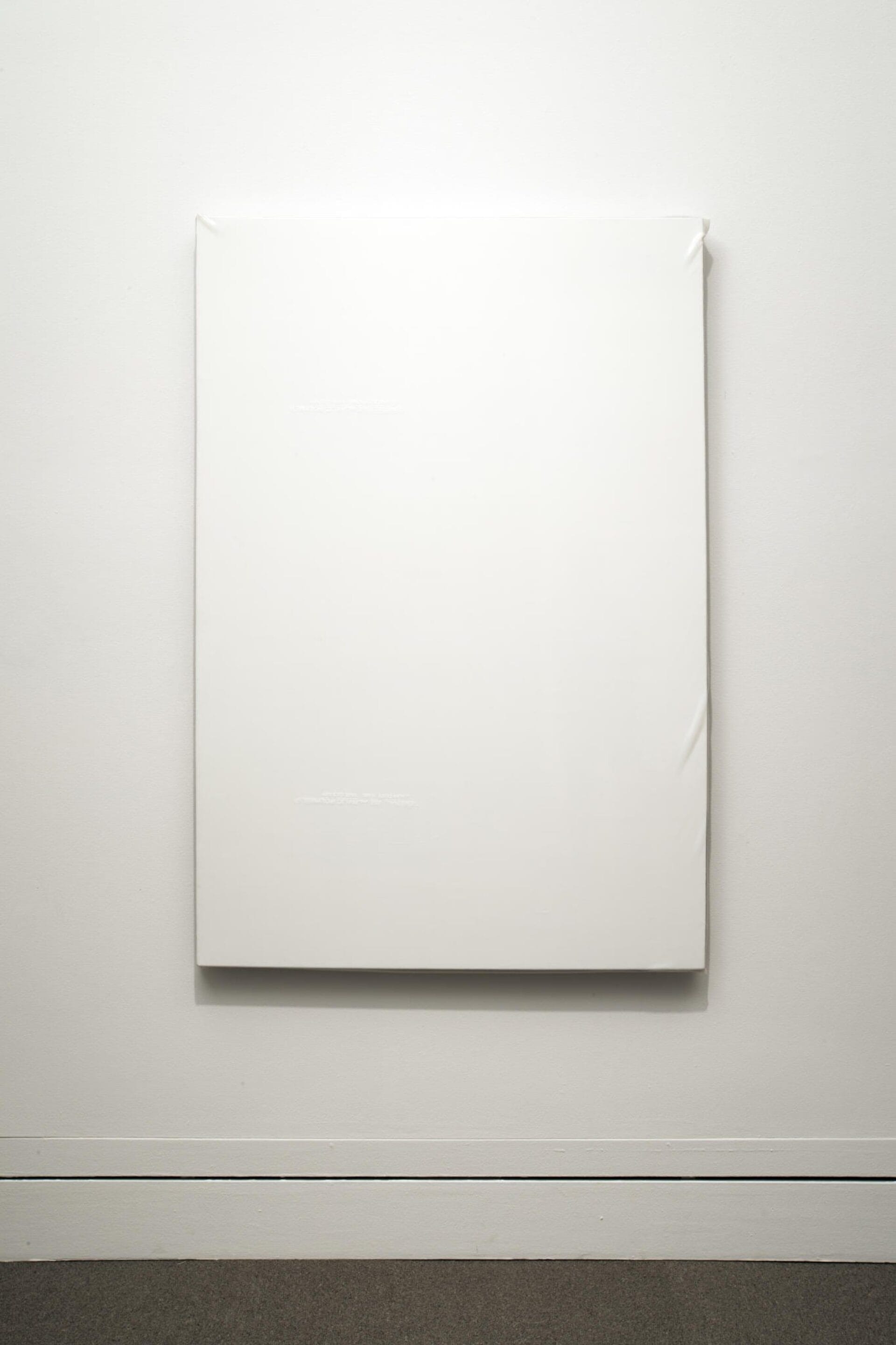 A minimalist art piece featuring a large, white rectangular canvas displayed on a plain white wall. The canvas appears to be blank, emphasizing simplicity and form. The floor below is light grey, creating a subtle contrast with the wall and canvas.