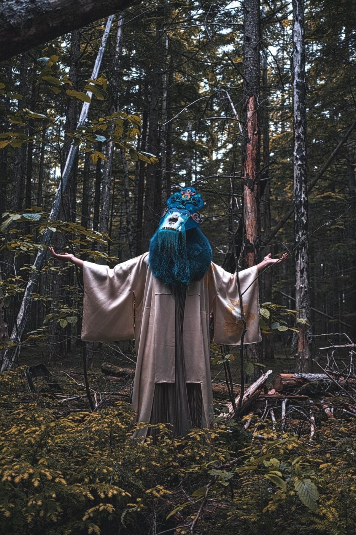 A person in a beige robe and a mask with blue fur and intricate designs extends their arms outward while standing in the dense forest of MacKenzie Gala. Various trees and greenery surround them, adding to the mystical atmosphere, as if drawn from SENSORIA's enchanting realms.