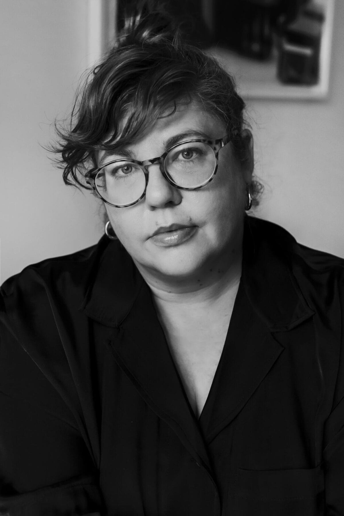 Black and white portrait of a person with wavy hair pulled back, wearing round glasses and a dark top. They have a neutral expression and are gazing slightly off-camera, reminiscent of how one might focus on a mesmerizing pinwheel. The background is blurred, making the person the focal point of the image.