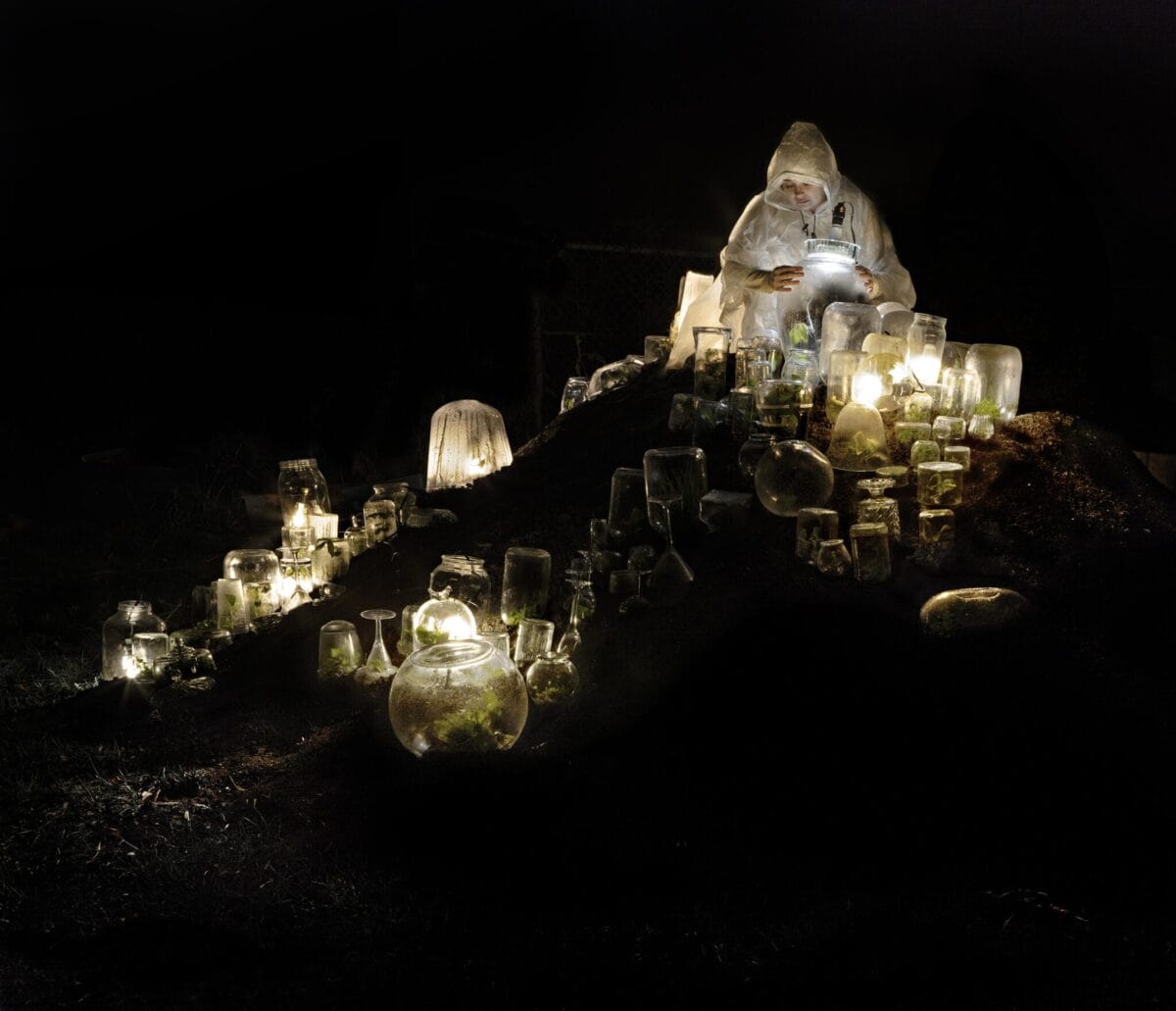 A person dressed in a white hooded outfit sits on a mound with many glass jars of various sizes glowing softly in the dark. The jars, reminiscent of SENSORIA's ethereal essence, contain plants and emit light, creating a mysterious atmosphere. The person is looking down, possibly at a jar.