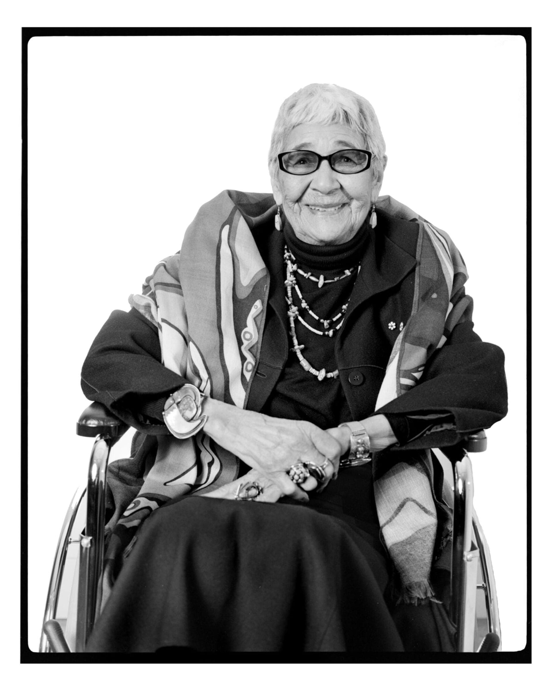Black-and-white photo of an elderly person sitting in a wheelchair. They are wearing dark clothing, a patterned shawl, several rings, and necklaces. They have short hair, wear glasses, and are smiling at the camera.