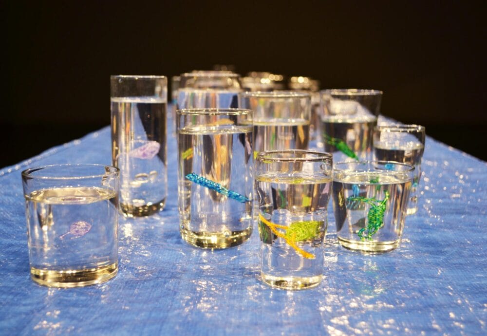 A collection of glass cups filled with water is placed on a blue surface. Each glass contains a small, colorful object resembling a sea creature, featuring various colors like green, blue, and yellow beads in the blood of the water.